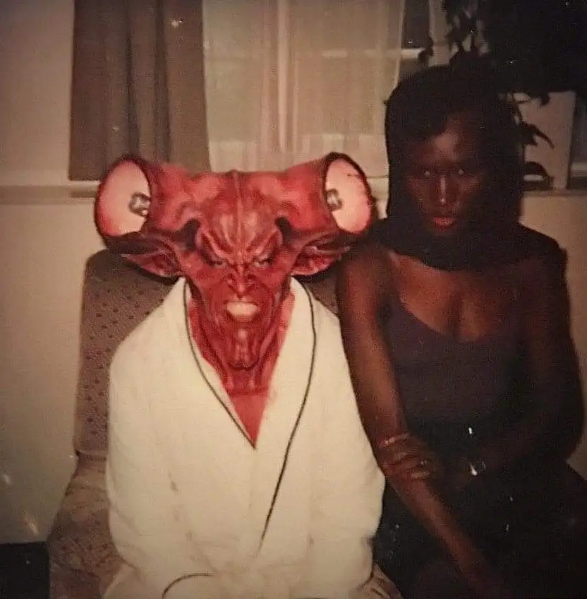 Tim Curry sat next to Grace Jones. He is wearing elaborate prosthetics and she is sat next to him wearing a black head scarf, dark spaghetti top and pants