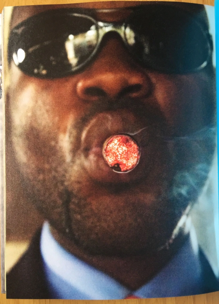 A Black Congolese man wearing sunglasses and smoking a cigar. He is wearing a suit (mostly out of shot)
